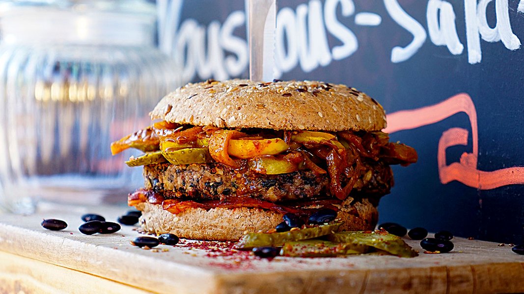 Auf einem Holzbrett ist ein veganer Burger angerichtet. Ein Black Bean Burger. Die Komponenten sind ein veganes Vollkornbun, der vegane Bohnen-Pattie und ein Apfel-Relish. In dem Burger steckt ein Messer
