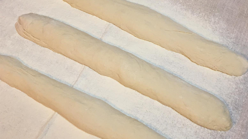 Baguette-Teiglinge die in der typischen Baguette-Form auf einem bemehlten Leinentuch liegen.